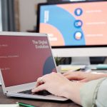 Profissional trabalha em um laptop em um escritório moderno, diante de uma tela com elementos visuais de evolução digital.