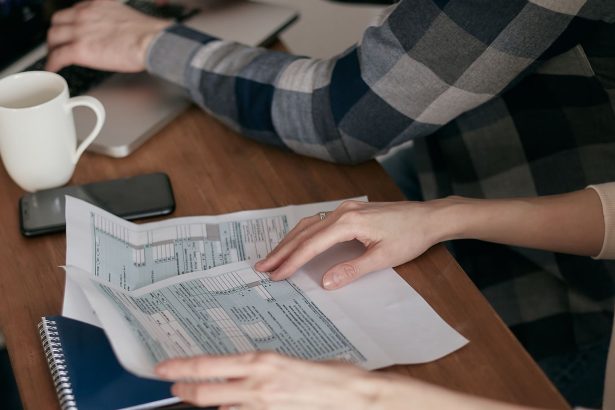 Mãos organizam formulários fiscais sobre uma mesa com calculadora, notebook e caderno.
