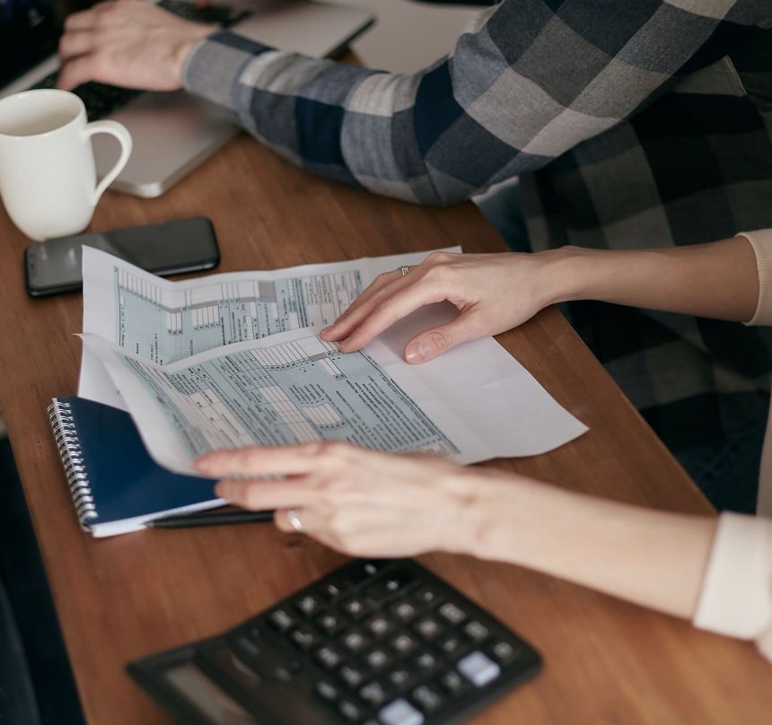 Mãos organizam formulários fiscais sobre uma mesa com calculadora, notebook e caderno.